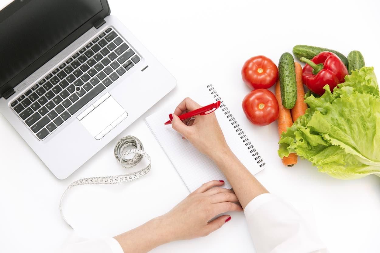 A woman writing on a writing pad with vegetables and a laptop near her.